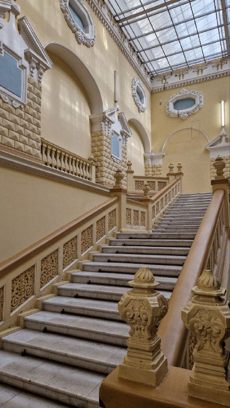 Широкая лестница с балюстрадами и декоративными перилами, детали неорусского декора и окна под куполом, интерьер в жёлто-белой гамме.