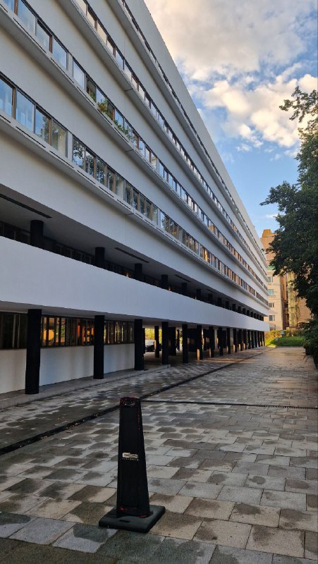 Длинный вид фасада Дома Наркомфина со стороны двора: продольная композиция, ряд опор и тёмные проходы под зданием, отражающие конструктивистскую эстетику.