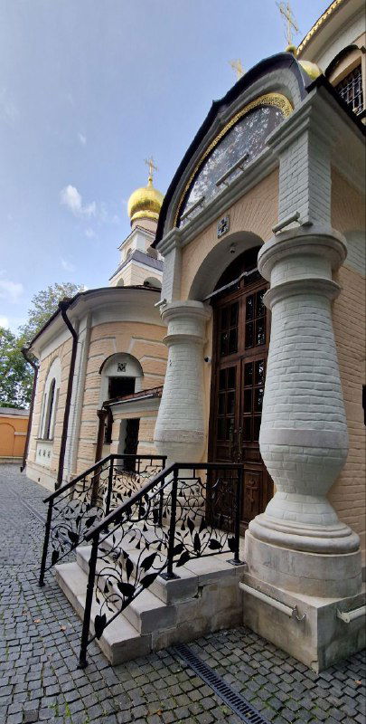 Фрагмент главного входа церкви: колонны, ступени и деревянные двери на фоне бело‑пастельного декора фасада и неба.