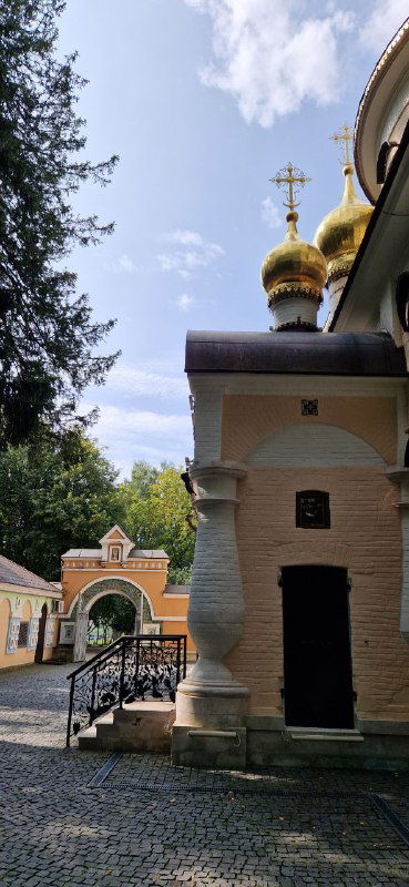 Вид на боковой корпус Спасо‑Преображенской церкви и прилегающую территорию с дорожкой, стенами пастельного цвета и небольшими постройками.