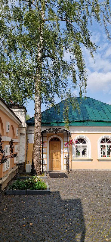 Небольшое пристроенное здание рядом с церковным двором, зелёная крыша, берёза на переднем плане и аккуратный двор у храма.