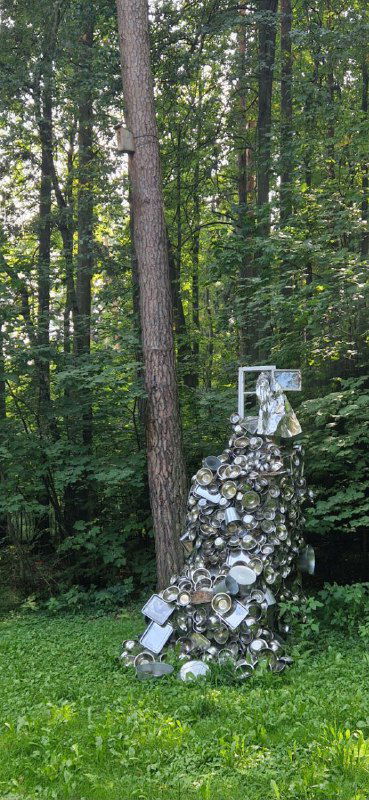 Скульптурная инсталляция у дома-музея: каменная колонна с вмонтированными предметами на фоне лесного массива и зелёной лужайки.