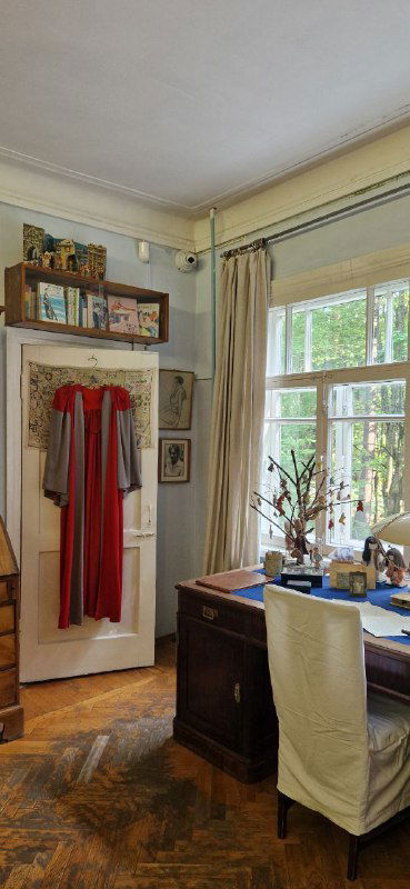 Рабочий кабинет писателя: стол с предметами, вешалка с мантийкой или одеждой и полки с сувенирами и детскими вещицами.