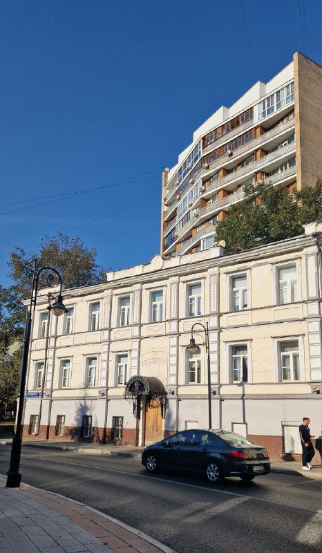 Городская улица со старинными домами на Спиридоновке: низкое историческое здание на фоне современного жилого корпуса, припаркованные машины и тротуар.