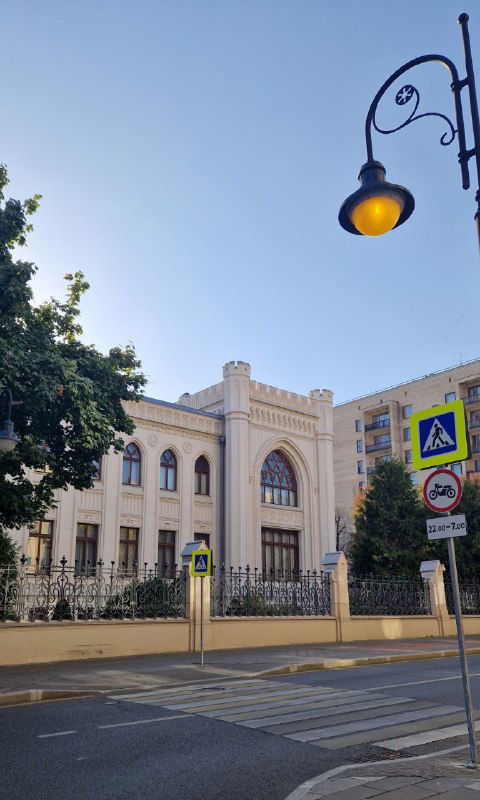 Вид через исторический уличный фонарь на белое официальное здание с высоким забором — одно из общественных сооружений на Спиридоновке, окружённое зеленью.