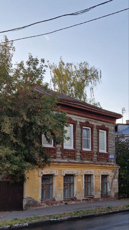 Традиционный деревянный дом в Серпухове с резными наличниками и слегка поцарапанной краской фасада, обрамлённый зеленью и ясным небом.