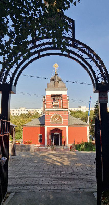 Вид через арочную металлическую ограду на краснокирпичный храм Коньково — входная зона с куполом и ухоженным двором, архитектура религиозного памятника.