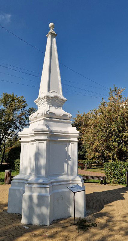 Белый обелиск-памятный столб в парковой зоне Коньково на фоне деревьев и голубого неба — исторический ориентир, упомянутый в тексте.
