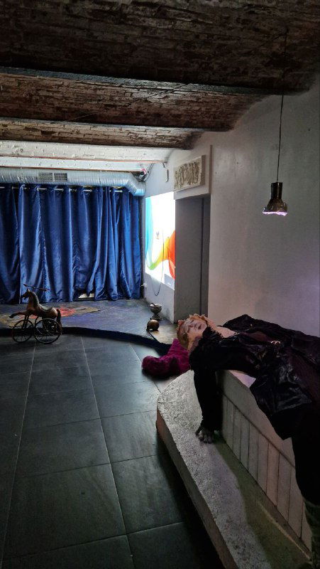 Интерьер клуба Лахесис перед спектаклем: низкий потолок с деревянными балками, синие шторы, ступени и посетитель на лавке — камерная атмосфера зала.