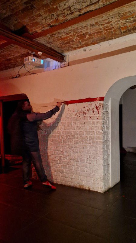 Технический кадр со сцены: художник или техник закрепляет декор на белой кирпичной стене клуба — момент подготовки или сценической смены.