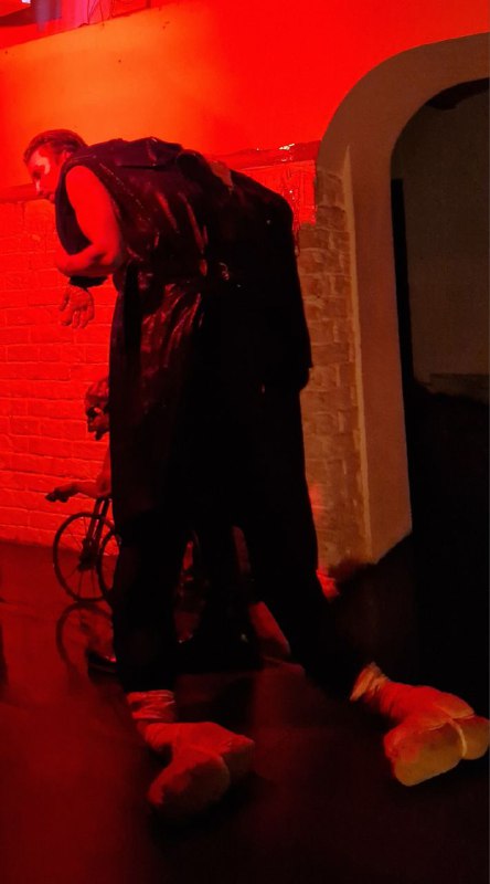 Интенсивный сценический момент: актёр наклоняется над сценой в тёмном костюме, виден реквизит на полу и сильный красный свет, создающий напряжение.