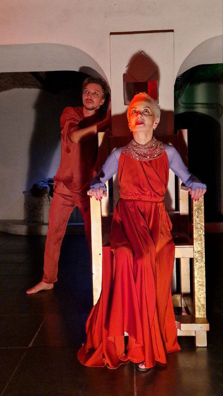 Сцена с актрисой на троне и партнёром рядом: костюмы, архитектурные ниши зала и контрастное световое решение подчёркивают драматизм номера.