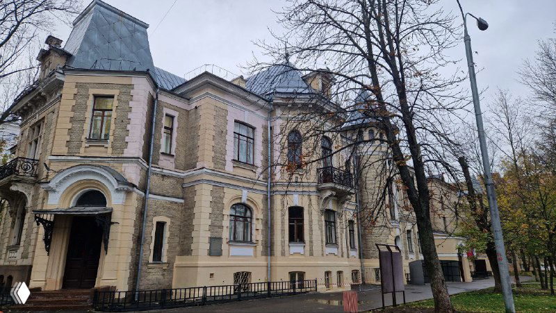 Панорамный снимок особняка в неоготическом/историческом стиле: многогранные объёмы, мансардные окна и аккуратная ограда на фоне осеннего неба.