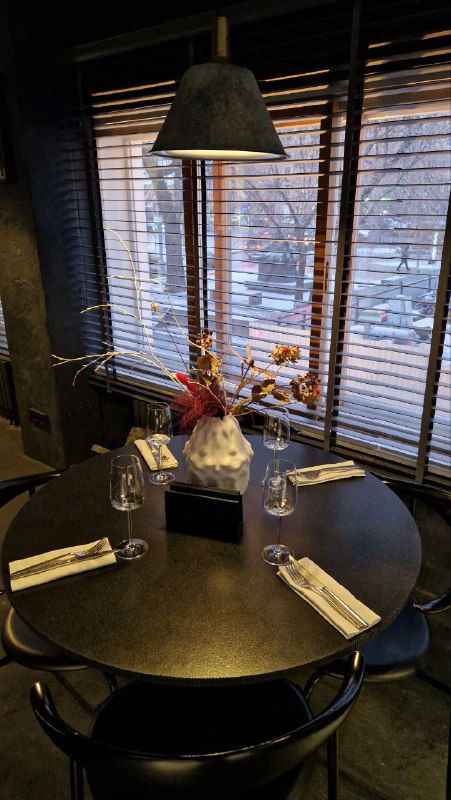 Ближний план застеленного круглого стола у окна: приборы, салфетки и вид на город через деревянные жалюзи с тёплым светом.