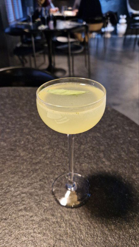 Крупный план пастельного коктейля в бокале на тёмной столешнице — прозрачный бокал, светлая жидкость и тонкая долька лимона.