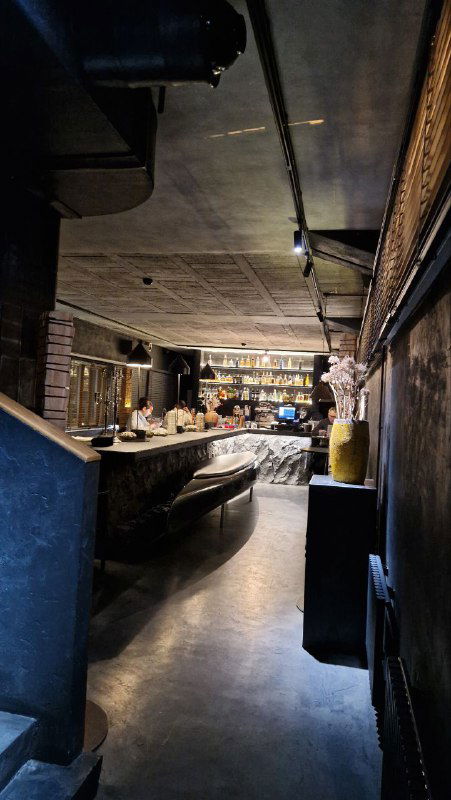 Барная зона и проход ресторана: длинная барная стойка, приглушённое освещение и интерьер в тёмных тонах с бетонным потолком.