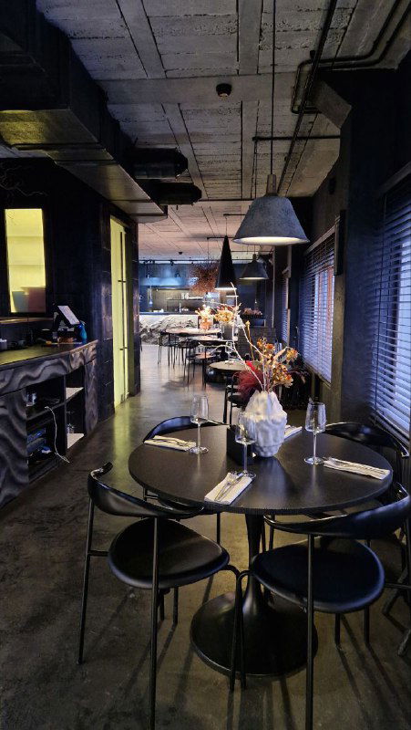 Ряд столов в темной зоне, большой напольный вазон со сухоцветами на переднем плане и мягкое освещение над столами ресторана.