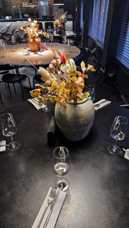 Крупный план декоративной вазы с сухими цветами на столе, окружённой бокалами и приборами — акцент на столовом оформлении и декоре.