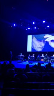 Сцена спектакля с крупной проекцией портрета на экране, ансамбль музыкантов слева и тёмный зал с публикой в переднем плане.