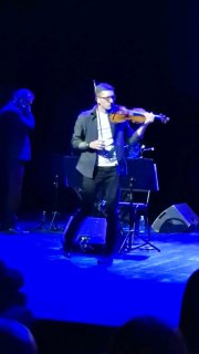 Антон Якушев играет на скрипке на сцене проекта САМИ: динамичное исполнение, видна поза скрипача и часть оркестра в синем свете.