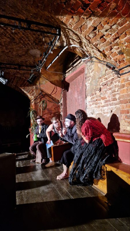 Группа актёров и зрителей на скамьях у стены в кирпичном подвале — ощущение близкого контакта и импровизационной интонации постановки.