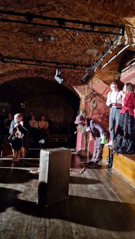 Широкий план сцены с несколькими актёрами: тёмный пол, кирпичный свод и сцены с вертикальными находками декора в постановке.