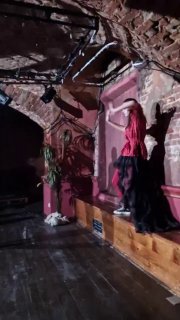 Вертикальная композиция: актриса в красном наряде стоит у стены, контраст с кирпичной фактурой и драматическим светом — эпизод спектакля.