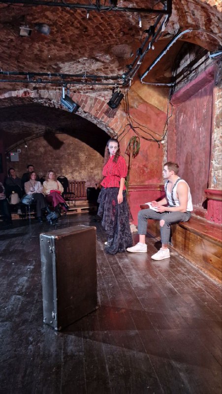 Два актёра на сцене — один в красном, другой в повседневной одежде; камерная сцена и прямой контакт с публикой создают эффект присутствия.