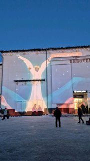 Проекция оживших детских рисунков на фасаде кинотеатра «Родина»: крупный светящийся ангел и детские сюжеты на стене вечером.