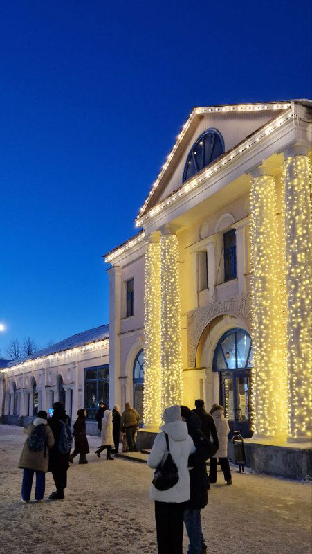 Историческое здание, украшенное гирляндами и огнями: фасад, прохожие и зимняя улица в вечернем освещении фестиваля.