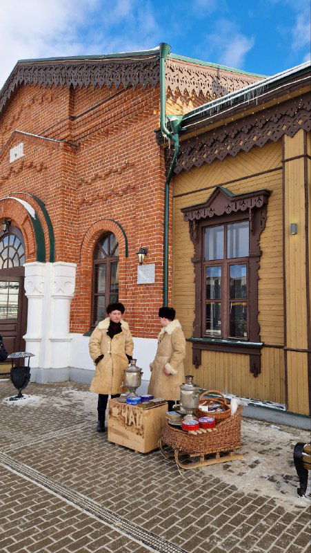 Уличные рождественские развалы и люди в тёплой одежде у вокзала: деревянные столы с атрибутикой и самоварами на снегу.