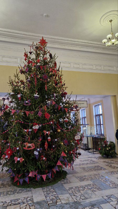 Украшенная рождественская ель внутри вокзального зала: высокий нарядный ёлочный декор, гирлянды и бумажные игрушки на фоне окон.