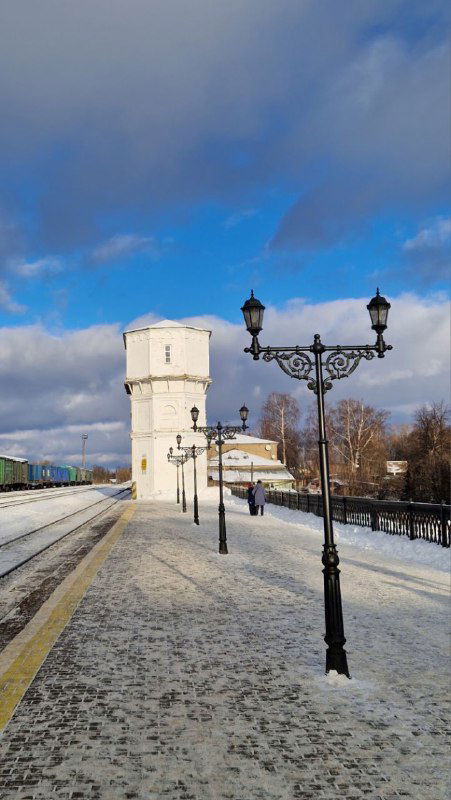 Платформа и белая башенка на фоне зимнего неба: заснеженная железнодорожная платформа, фонари и перспектива путей.