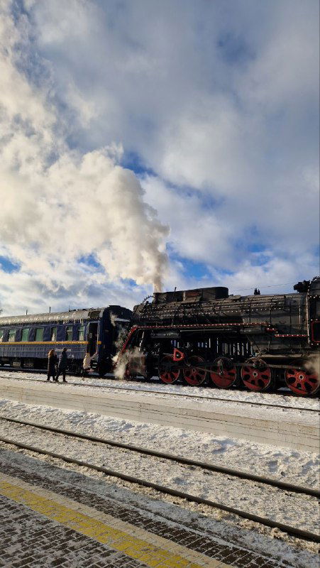 Старинный паровоз, украшенный Вифлиемской звездой, с паром и ретро-вагонами на станции — рождественский экспресс на путях.