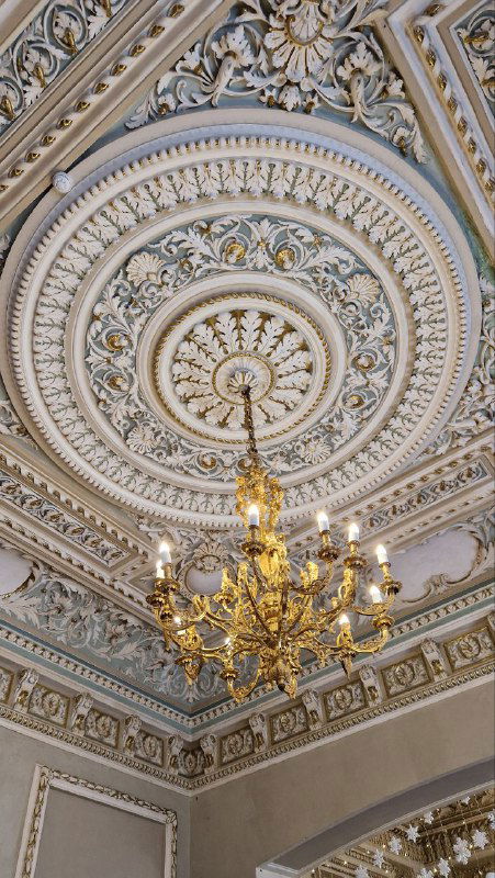 Детальная фотография лепного потолка с розеткой и хрустальной люстрой в одном из залов усадьбы М.А. Павлова; видно сложную декоративную лепнину и золочение.