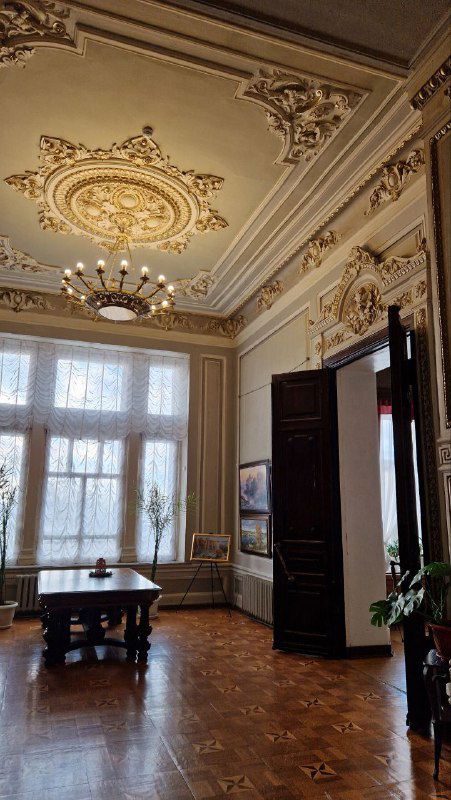 Парадная гостиная усадьбы с высокими окнами, лепным декором на потолке и деревянным паркетом; частично отреставрированный интерьер XIX века.