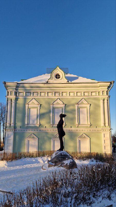 Историческое здание с фасадом пастельного цвета и скульптурой на постаменте на фоне чистого синего неба в Шуе.