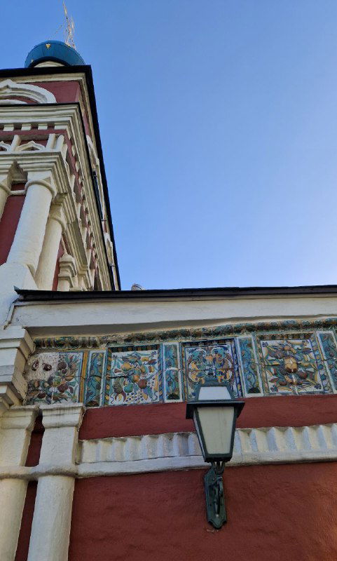 Крупный план узорных керамических изразцов и лепнины на стене храма, декоративный фриз вблизи уличного фонаря.