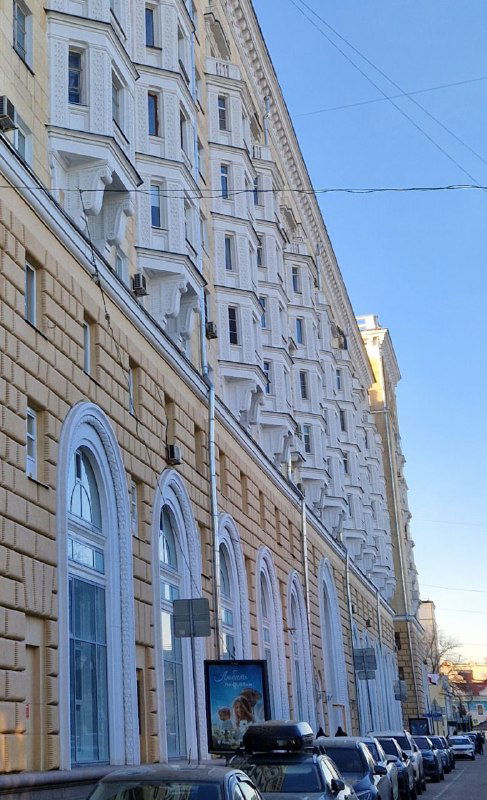 Протяжённый фасад доходного дома с арочными и прямоугольными окнами, архитектурные детали вдоль тротуара и парковки.