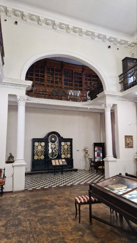 Общий вид зала музея с астрономическими часами на дальней стене: колонны, балкон, витрины и атмосферный классический интерьер экспозиции в Иваново.