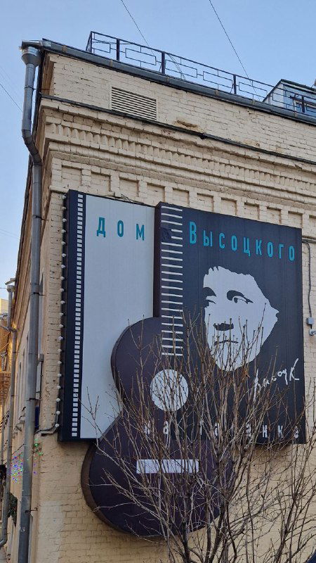 Билборд с изображением Владимира Высоцкого в виде гитары на фасаде музея: крупный портрет, уличный пейзаж и детали городской среды вокруг.
