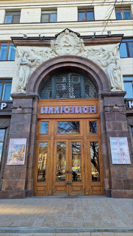 Парадный деревянный вход в кинотеатр с резными дверями и надписью 'Иллюзион', отражающими историческую атмосферу места и кинематографическую тематику.