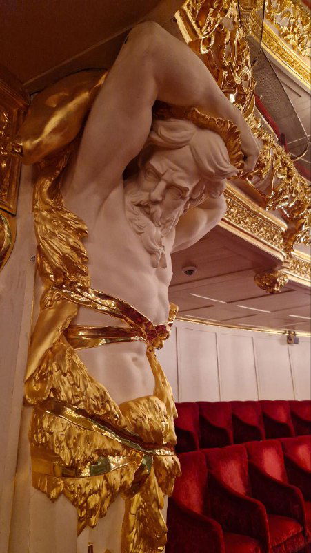 Деталь позолоченной скульптуры-атланта и фрагмент ложи с бархатными креслами и золочёными элементами театрального декора.