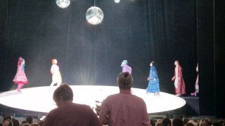 Перспектива из партера: несколько актёров в разноцветных костюмах идут по круглой сцене; передний план — зрители, подчёркивающие интерактив постановки.