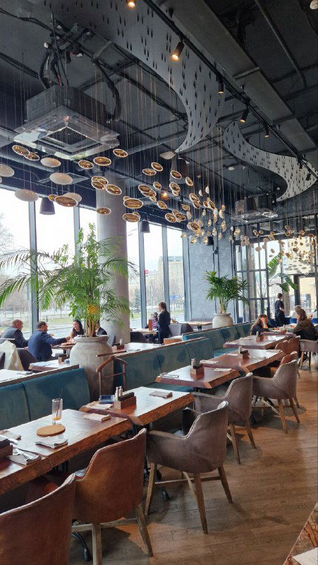 Широкий зал с длинными рядами столов у панорамных окон и оригинальными подвесными светильниками, вид на посадочные места и дневной свет в ресторане.