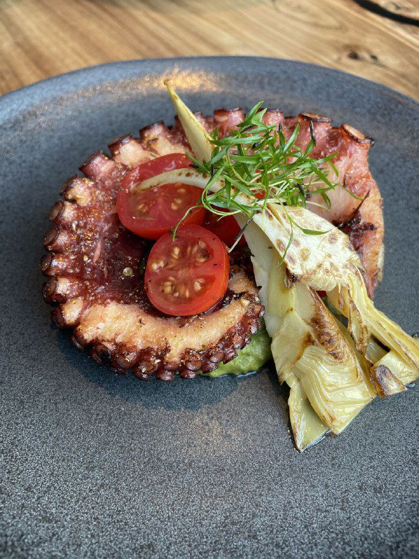 Блюдо с обжаренным осьминогом, украшенным помидорами черри и зеленью, подача на тёмной тарелке — морской акцент в гастрономическом стиле ресторана.