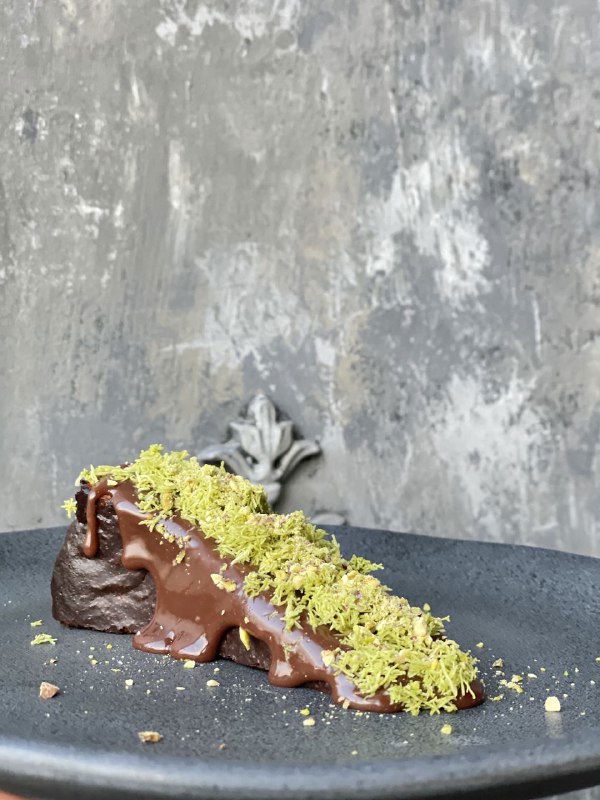 Шоколадный кусок десерта с зелёной крошкой на тёмной тарелке — концептуальный чизкейк «дубайский шоколад», текстура и контраст цветов в кадре.