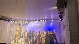 Актёры на сцене небольшого белого зала Театра «МОСТ»: ансамбль в светлых костюмах, направленный сценический свет и минималистичные декорации, зрители в первом ряду.