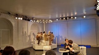 Сцена спектакля «ГУДОК!»: актёры за столом в интерьере редакции, приглушённые тона освещения и архитектурные арки на заднике спектакля.