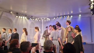 Групповой выход актёров на поклон: сцена заполнена персонажами, эмоциональные позы, направленные прожекторы и лица зрителей на переднем плане.